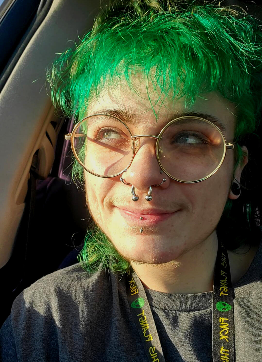 Jasper Hopkins Image of a person with round glasses, green hair, and piercings smiling with the sun hitting their face.